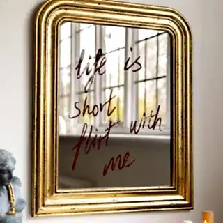 A gold mirror with the words 'life is short flirt with me' written on it, styled on a wall above some lit candles and an ornament.