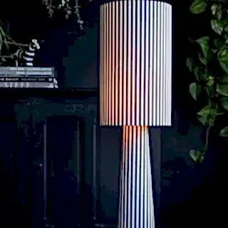 A black and white stripe floor lamp in front of a dark background with greenery.
