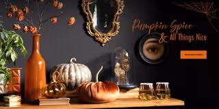 A styled Halloween-themed tabletop scene with orange and gold pumpkins, a tall amber vase with dried flowers, a glass cloche, two glasses of amber drink, and decorative wall art against a dark wall. Text on the right reads 'Pumpkin Spice & All Things Nice' with a 'Shop Halloween' button.