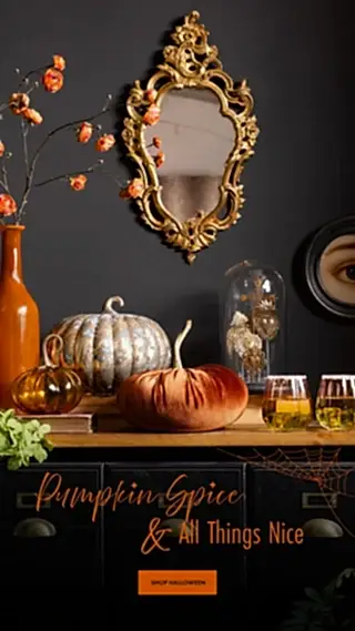 A styled Halloween scene with a dark wall and console table. A large ornate gold mirror hangs above the table, alongside a tall orange vase with dried orange flowers, a white ribbed pumpkin, a rust-colored pumpkin, a glass cloche, and three glasses of amber liquid.