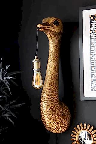 The Antique Bronze Ostrich Wall Light on a black wall, styled with a faux plant, framed artwork and mirror.