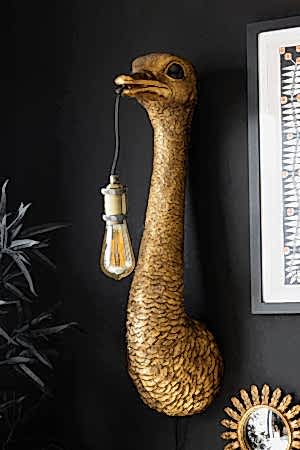 The Antique Bronze Ostrich Wall Light on a black wall, styled with a faux plant, framed artwork and mirror.