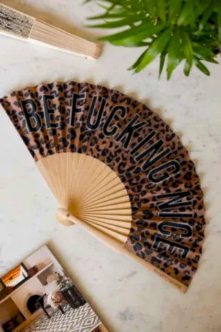 The Be Fucking Nice Leopard Print Fan displayed on a white marble table with another fan, magazine and a plant.