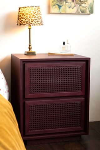 The Burgundy Rattan Bedside Table styled next to a bed with a switched on lamp on the top.