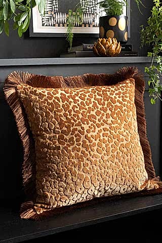 The Ochre Leopard Velvet Cushion with Fringe Trim styled on a black bench with some plants and accessories in the background.