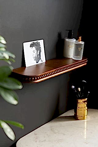 The Dark Wood Bobbin Floating Shelf  shown against a black wall with beauty products and a post card on the shelf. 