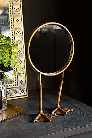 Gold Bird Legs Tabletop Mirror shwon against black walls with gold accessories. 