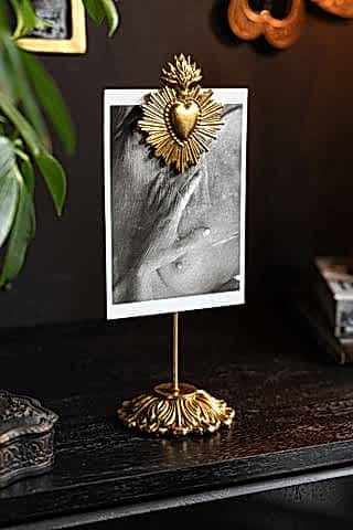 Gold Milagro Heart Photo Stand shown on a black sideboard with a black and white postcard on display. 