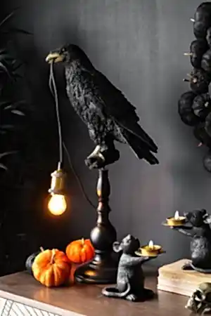 Black crow lamp on a sideboard styled with orange velvet mini pumpkins, mice candles and a skull ornament. The crow lamp is lit and the setting is giving Halloween vibes.