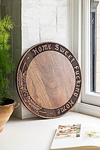 The Home Sweet Fucking Home Wooden Serving Board shown in a kitchen by the window against white zellige-style tiles. 