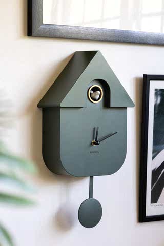 Beautiful khaki green cuckoo clock with a white cuckoo. The clock is hung on the wall with art prints in the background.