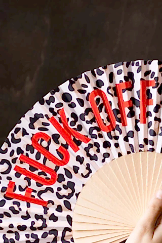 Close-up of the Leopard Print 'Fuck Off' Wooden Hand Held Fan in front of a dark wooden wall being held by a model's hand.