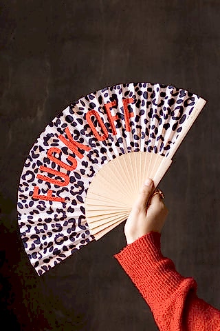 The Leopard Print 'Fuck Off' Wooden Hand Held Fan being held by a model in front of a dark brown wall.