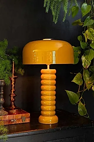 Lifestyle image of the Modern Metal Mustard Table Lamp displayed on a black sideboard surrounded by various other home accessories and plants.