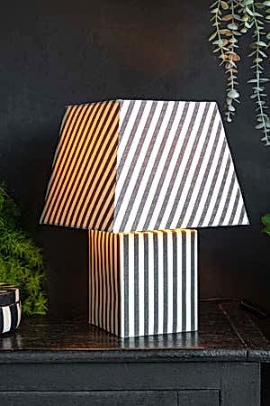 The Monochrome Stripe Table Lamp switched on and displayed on a black sideboard with some plants.