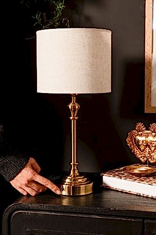 Person turning on a brass table lamp with a white shade on a dark surface.