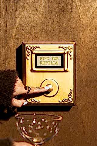 The Ring For Refills Gold Wall Ornament shown against a wood wall with someone holding up a champagne glass and pressing the button.