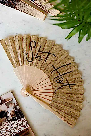 The Shit Hot Black & Gold Glitter Fan displayed on a marble table other fans, a magazine and a plant.